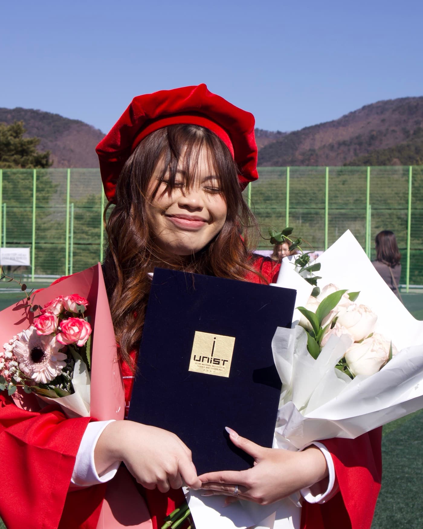 Gail Parayno at her UNIST graduation in Ulsan, in the red gown and cap, holding her diploma and two bouquets